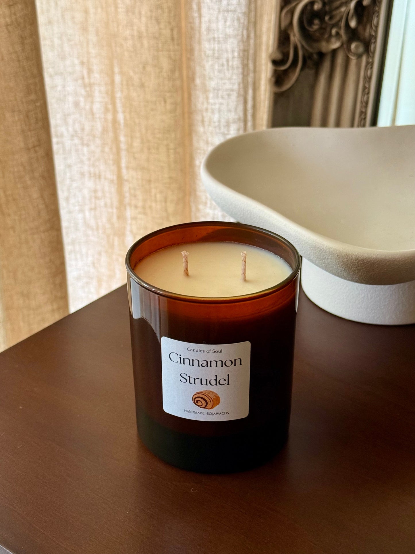 Braune Duftkerze mit dem Label „Cinnamon Strudel“ steht auf einem Holzregal neben einer dekorativen weißen Schale; im Hintergrund ein beiger Vorhang.