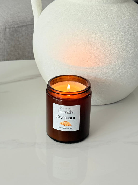 Braune Glasduftkerze mit dem Label "French Croissant" brennt auf einer weißen Marmoroberfläche neben einer weißen, strukturierten Vase – die Kerze vermittelt eine gemütliche, moderne Atmosphäre mit französischem Flair.