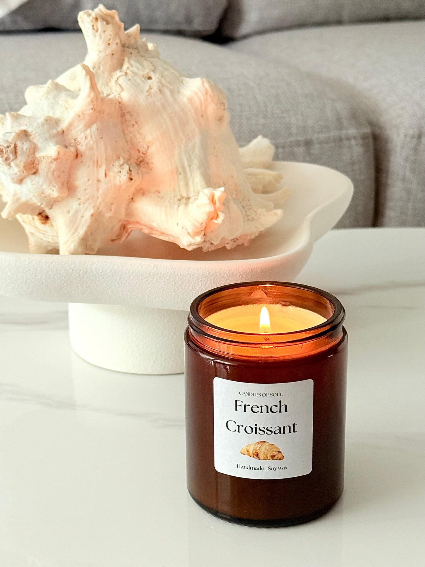 Duftkerze im Glas mit dem Aroma von französischem Croissant, auf einem hellen Tisch mit Frühstücksdekoration arrangiert.