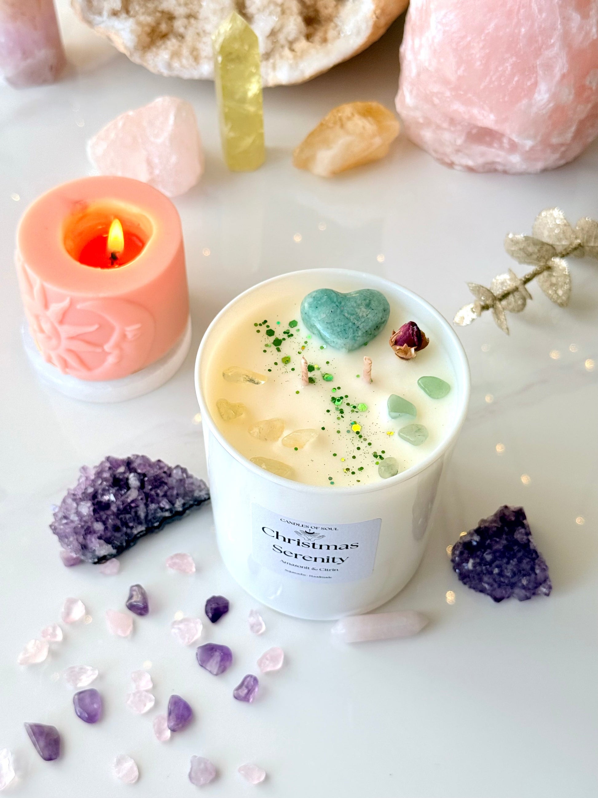 Weiße Sojawachs Duftkerze im Glas mit Label Christmas Serenity, dekoriert mit grünem Amazonit Herz und Citrin Splittern, im Hintergrund weihnachtliche Lichter und Kristalle