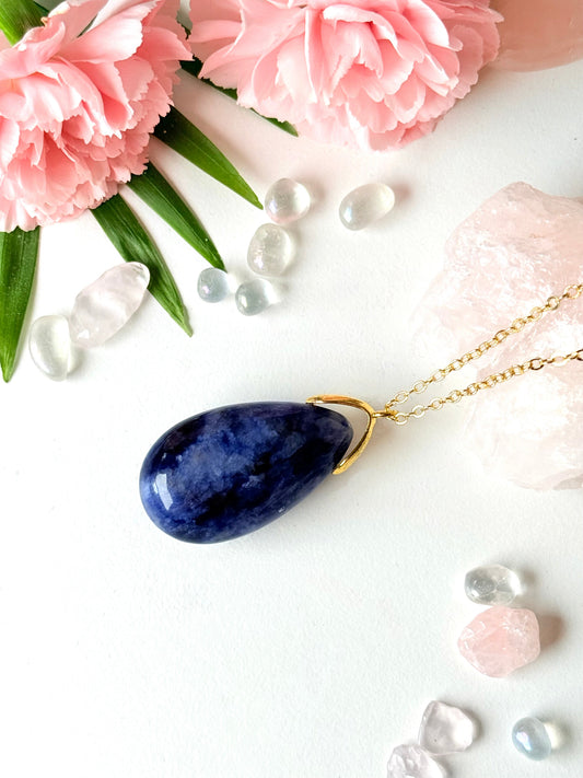 Blauer Sodalith-Tropfenanhänger mit goldfarbener Kette auf rosa Kristallen