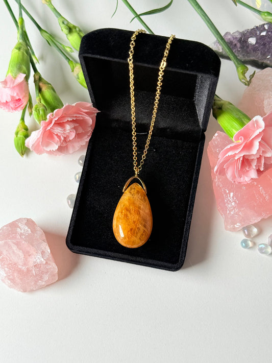 Handgefertigter Eisenquarz Tropfenanhänger auf goldener Kette, umgeben von rosa Nelken und Edelstein-Dekoration
