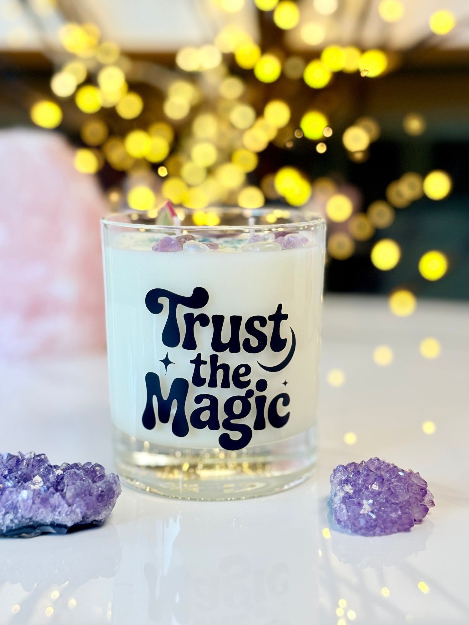 Handgemachte Sojawachskerze im klaren Glas mit dem schwarzen Schriftzug Trust the Magic, dekoriert mit Amethyst-Kristallen, einer getrockneten Rosenknospe und blauem Glitzer.