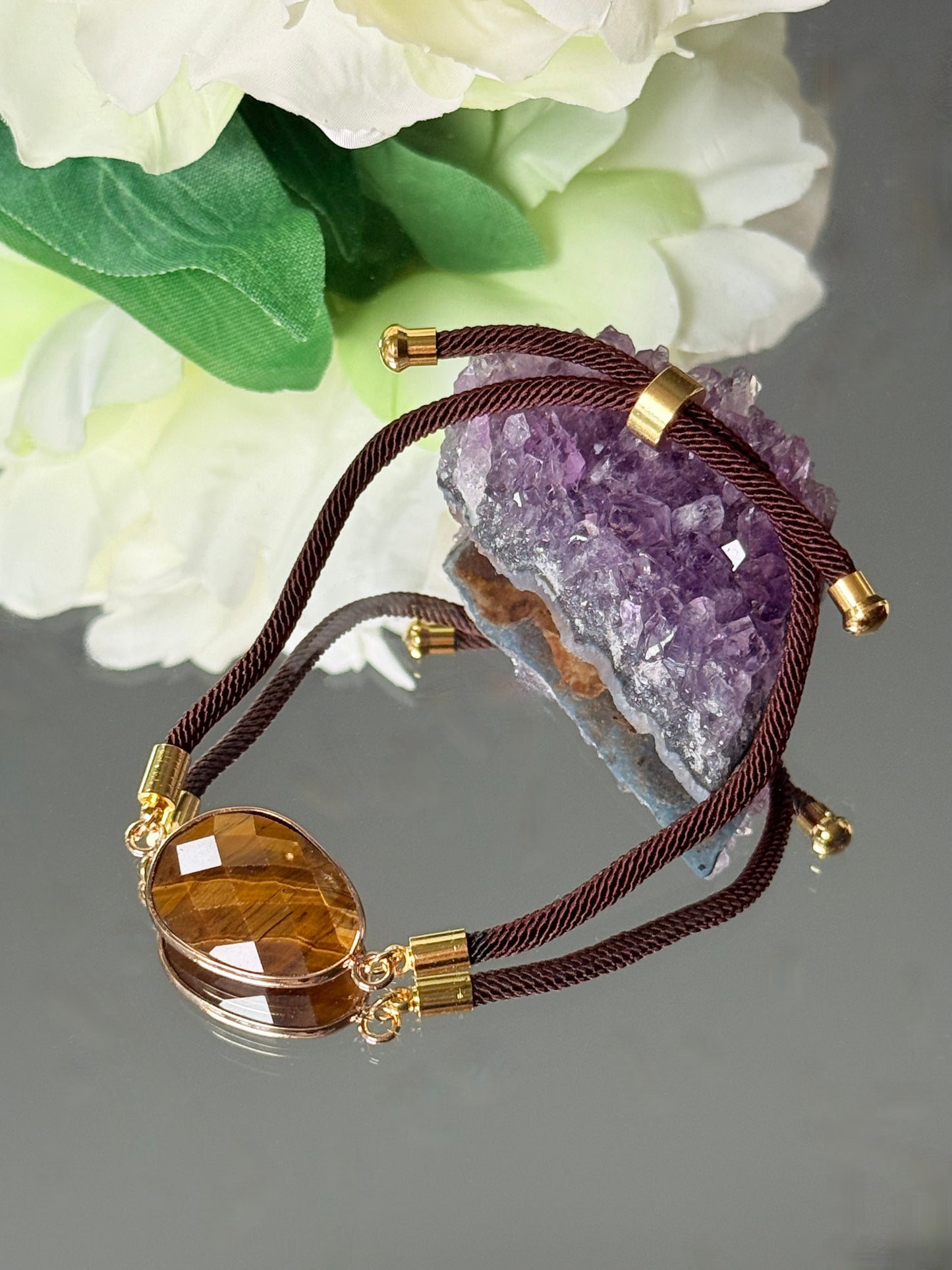Spirituelles Schutzarmband aus Tigerauge, dekorativ platziert auf einer spiegelnden Fläche neben Amethyst und weißen Blüten.