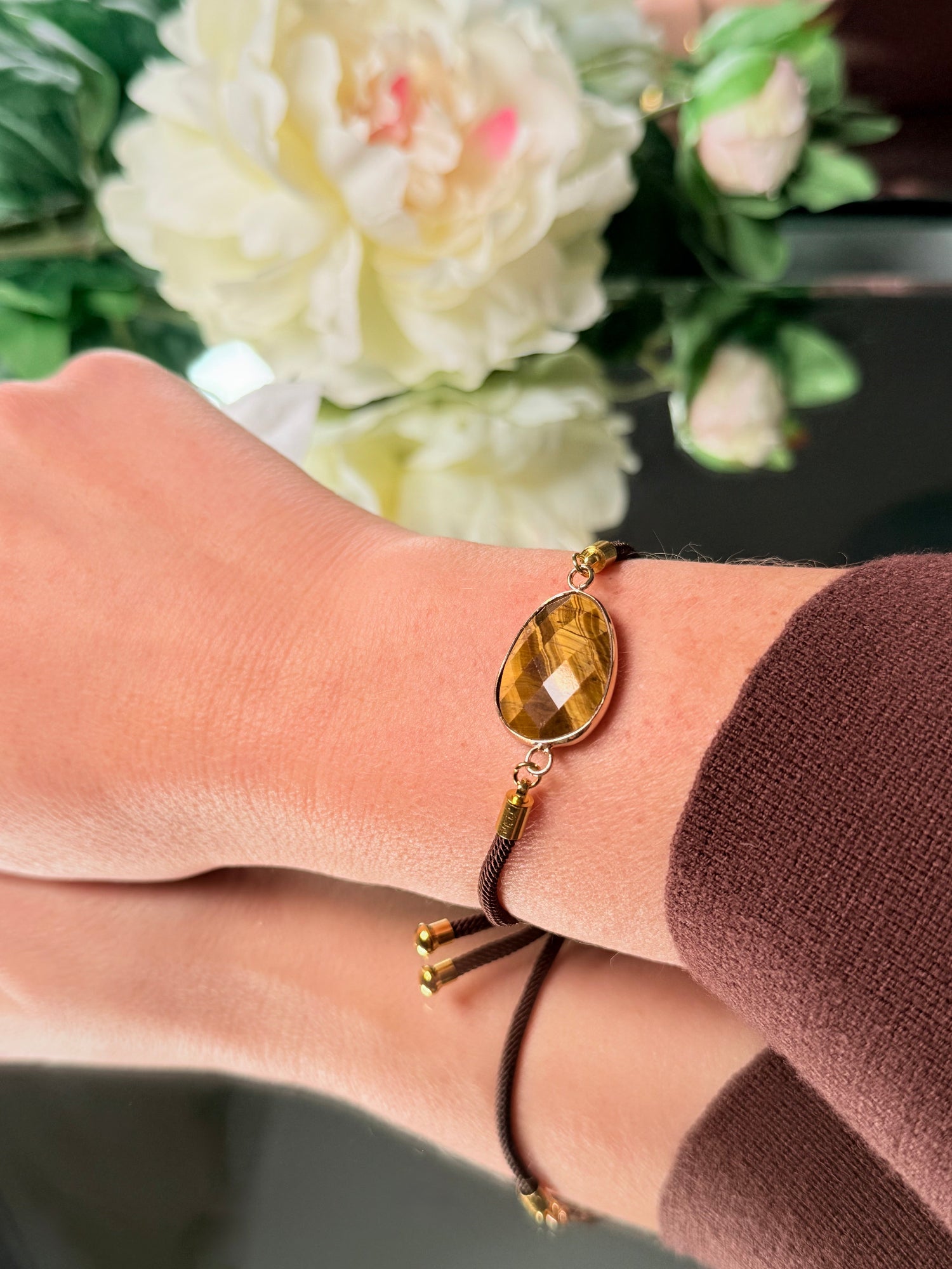 Nahaufnahme eines facettierten Tigerauge-Steins in Tropfenform, gefasst in ein braunes Kordelarmband mit goldenen Endstücken.