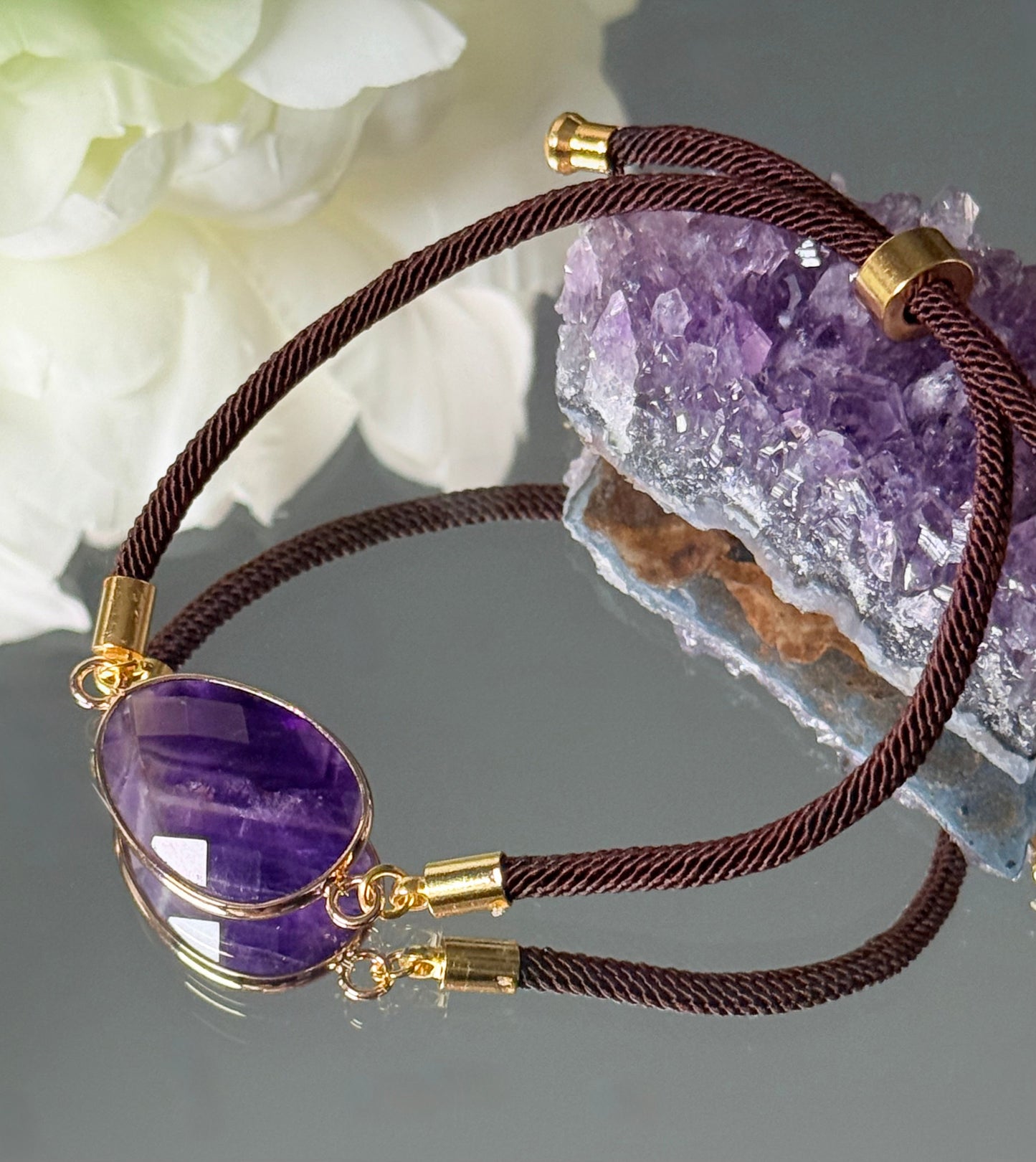 erstellbares Amethyst-Armband, stimmungsvoll arrangiert vor einem lila Rohkristall und weißen Blüten.