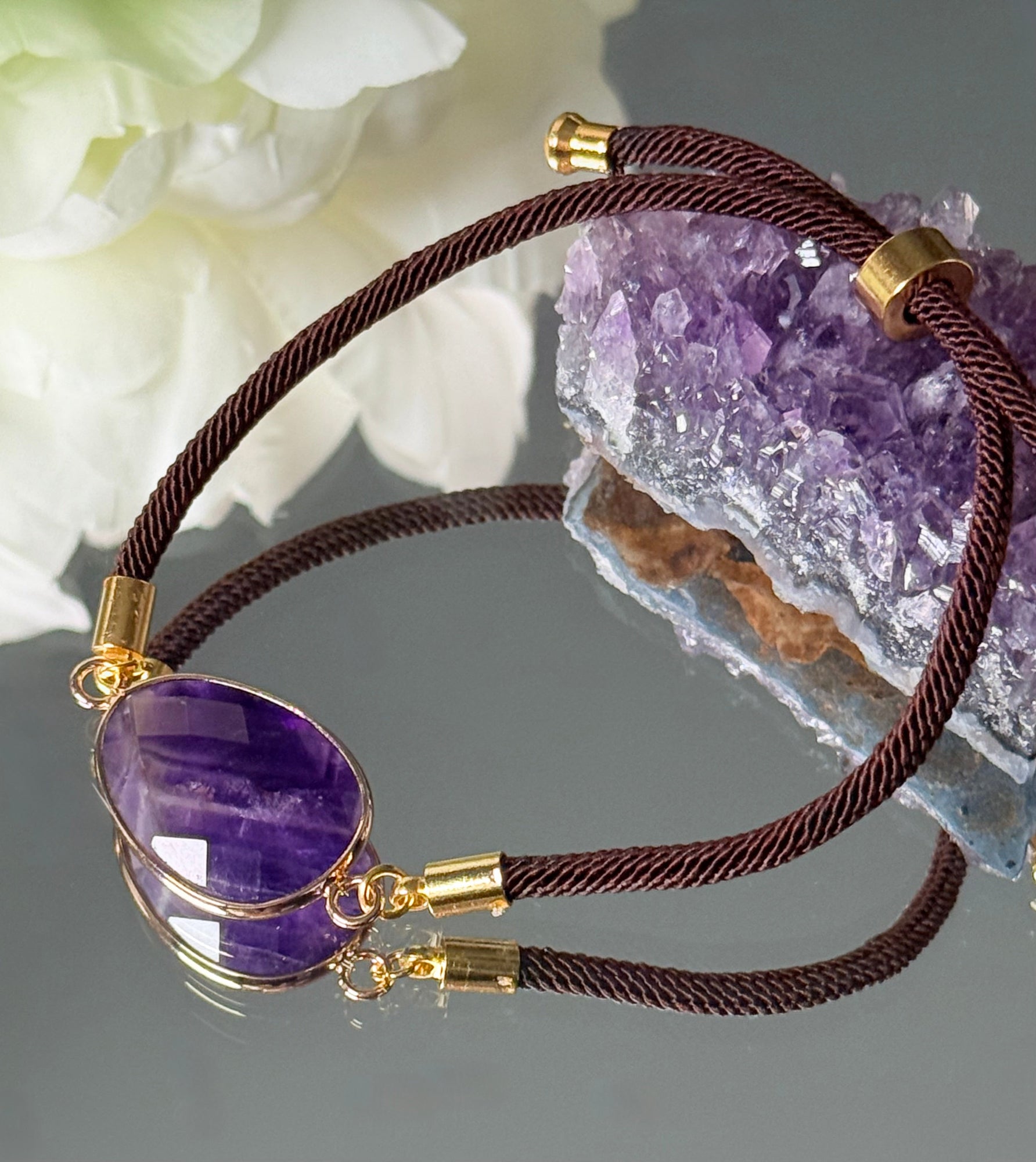 erstellbares Amethyst-Armband, stimmungsvoll arrangiert vor einem lila Rohkristall und weißen Blüten.