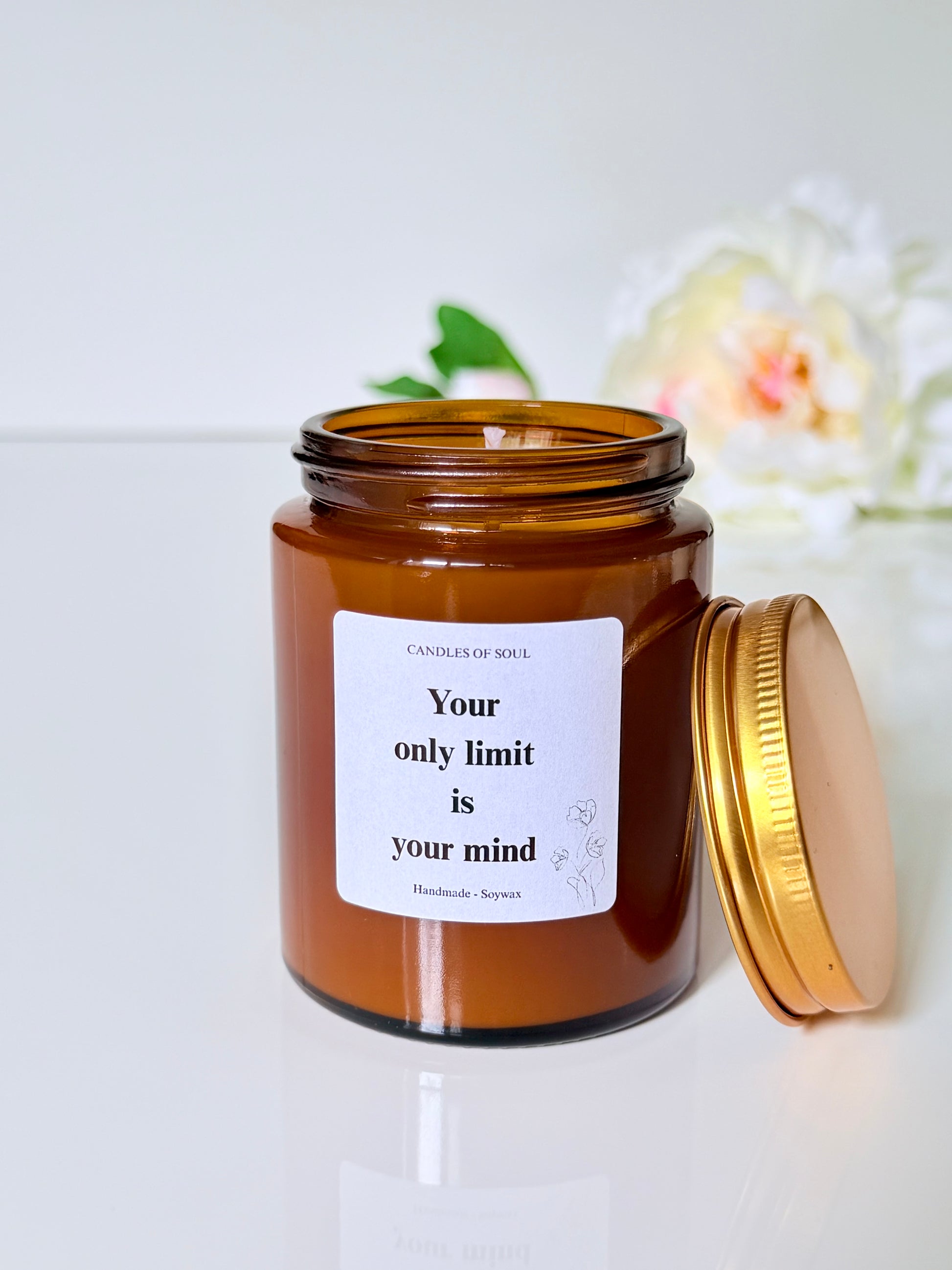 Geöffnete Motivationskerze aus Sojawachs mit goldenem Deckel neben dem braunen Kerzenglas vor einem hellen floralen Hintergrund.